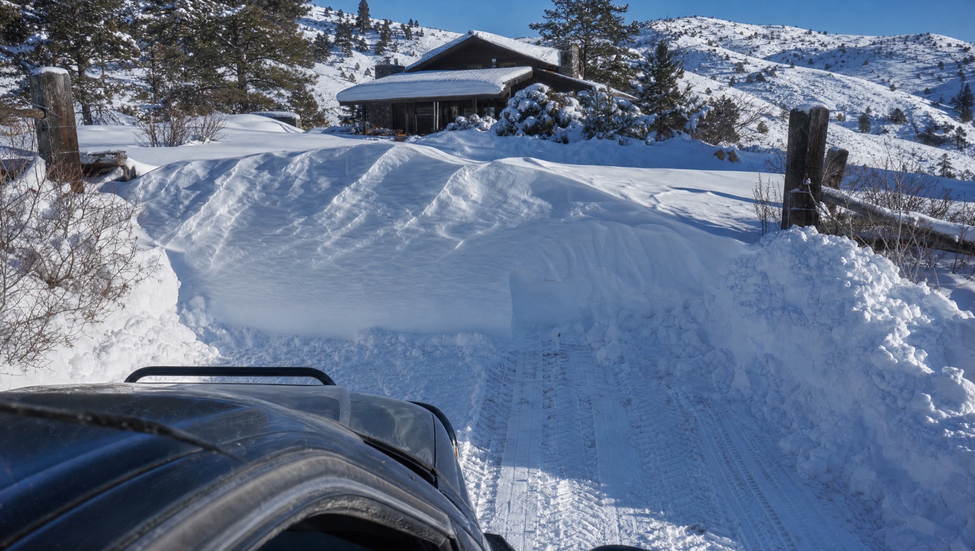 Tire tracks in packed snow stop dead at a large wind-carved drift completely blocking a private cabin driveway in Island Park, under a clear blue sky, with the cabin unreachable beyond it.